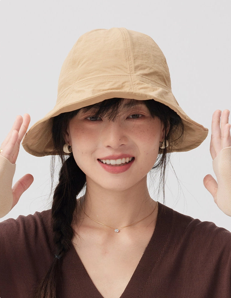 Front view of a woman smiling in a khaki wide brim bucket hat with both hands raised beside the brim.