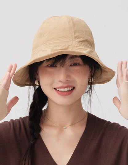 Front view of a woman smiling in a khaki wide brim bucket hat with both hands raised beside the brim.