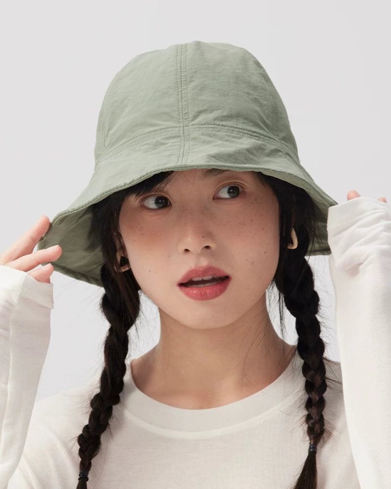 Woman holding the sides of a sage green bucket hat's wide brim while looking upward.