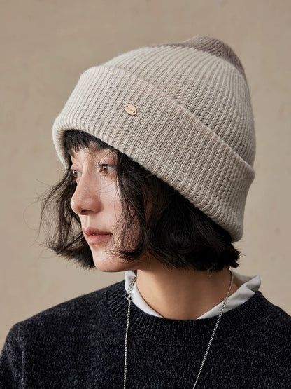 Side view of a model wearing a beige taupe wool color block beanie with a wide ribbed cuff and gold-tone detail.