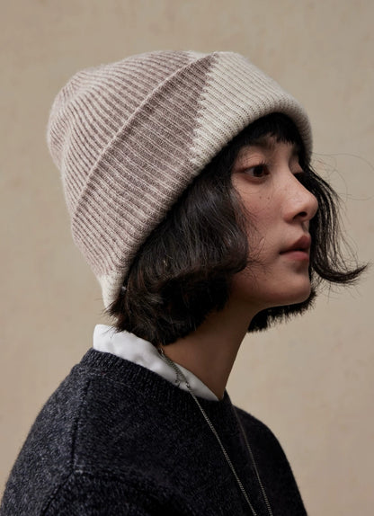 Right-facing profile of a model wearing a beige taupe wool color block beanie with diagonal panels and a folded cuff.