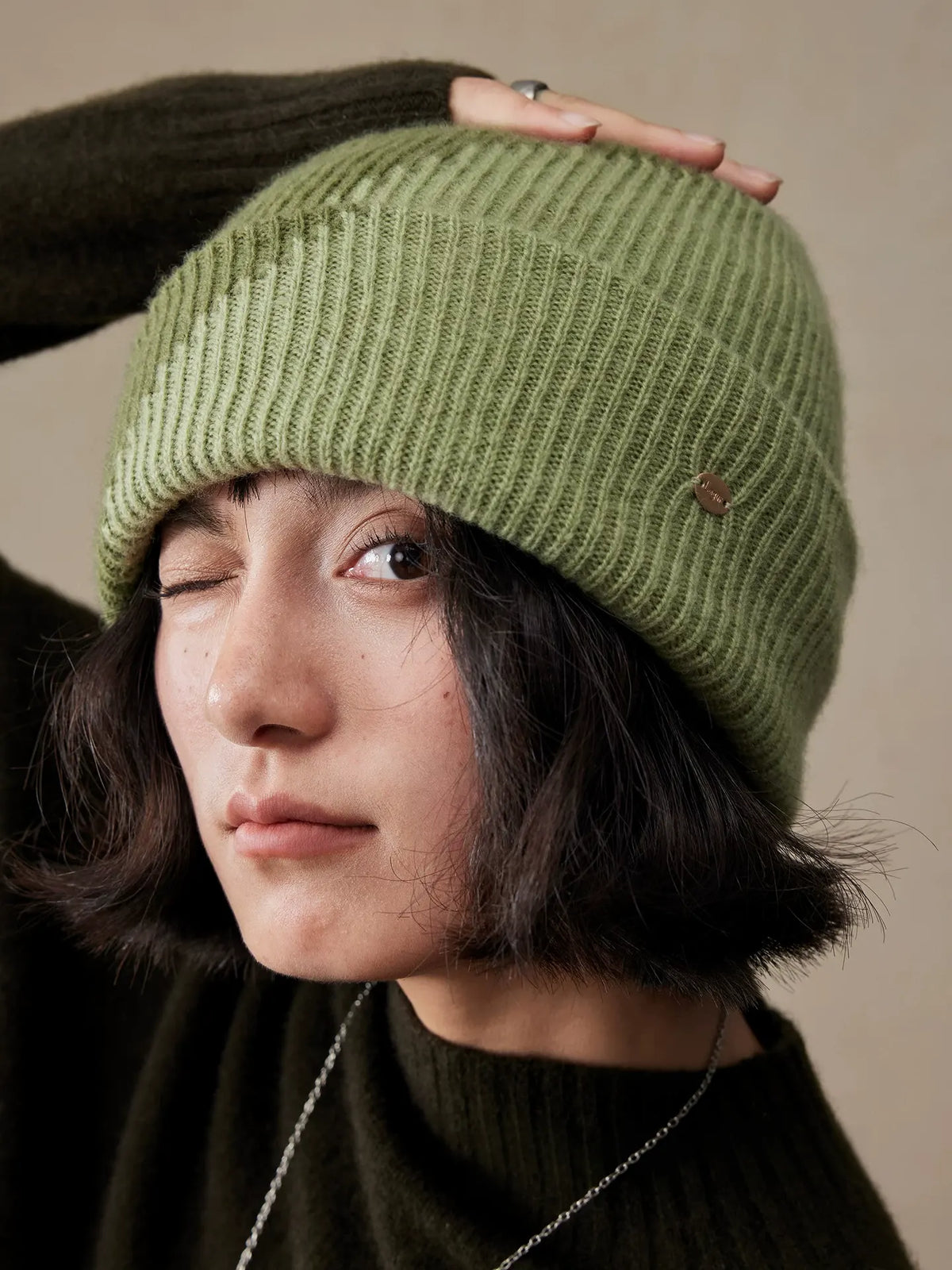 Model resting a hand on the crown of a moss green wool color block beanie, showing the ribbed knit and wide cuff.