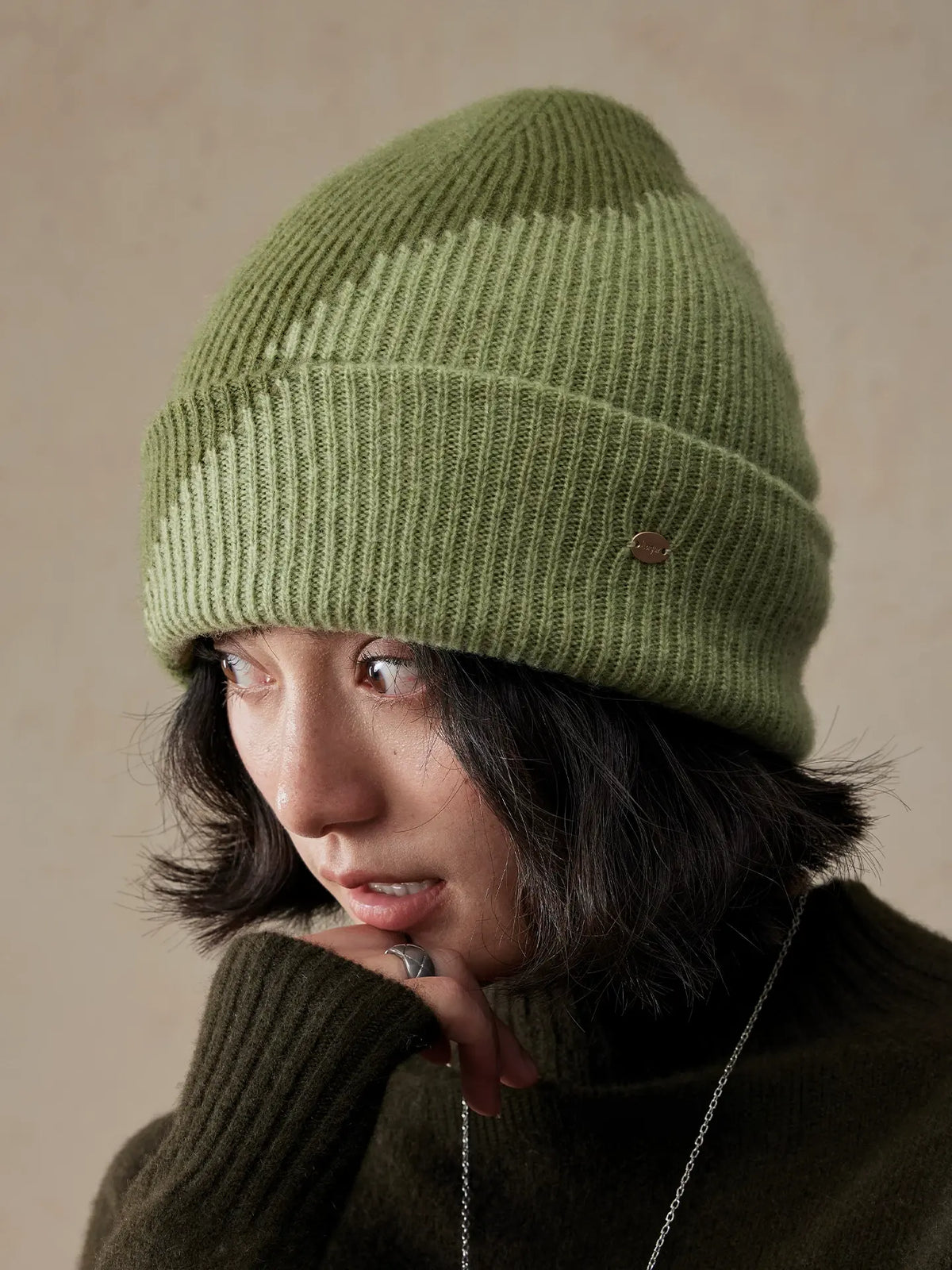 Model looking down in a moss green two-tone wool beanie, with a hand near the chin and the ribbed cuff visible.