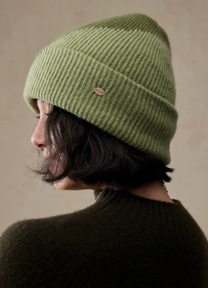 Rear three-quarter view of a model in a moss green wool color block beanie with a wide ribbed cuff and gold-tone detail.